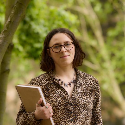 A photograph of Holly holding a sketchbook with trees in the background 